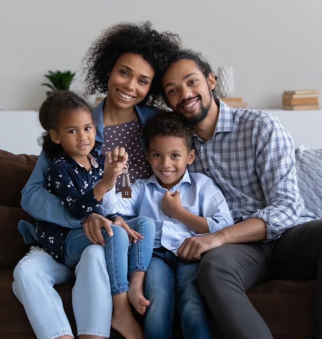 A black husband and wife wtih their young daughter and son sitting on a couch all smiling because they purchased a new home.