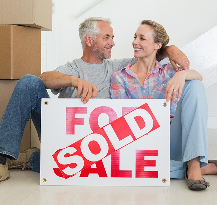 A happy middle aged white couple smiling holding a for sale sign with a sold sticker on top as they have evidentally just sold their home.