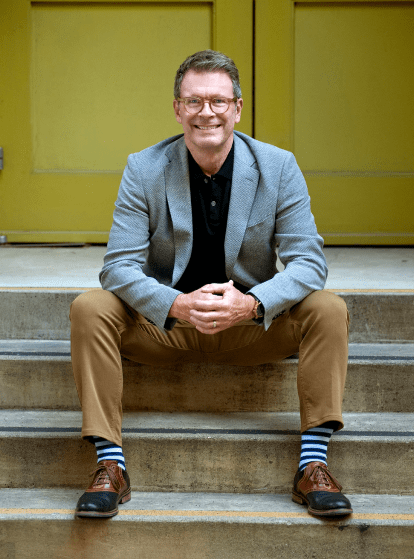 Realtor Tim DeBellis of Sonoma County California smiling while sitting on steps wearing a gray sports jacket, tan pants, and stripped socks.
