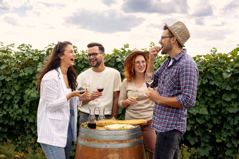 Two 20 something men and two 20 something women standing in a vineyard around a wine barrel enjoying a glass of red wine.